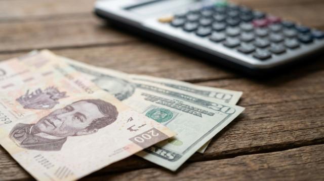 Primer plano de billetes de pesos mexicanos y dólares estadounidenses sobre una mesa de madera con una calculadora financiera de fondo, iluminación profesional