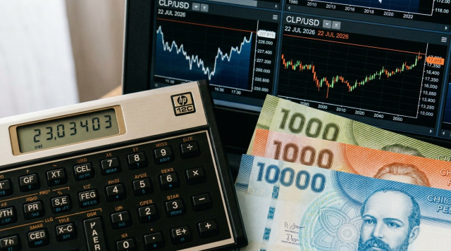 Primer plano de una calculadora financiera junto a billetes de mil y diez mil pesos chilenos y una pantalla con gráficos de divisas