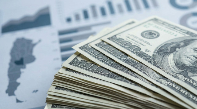 Primer plano de billetes de cien dólares estadounidenses sobre un fondo con gráficos financieros y el mapa de Argentina en tonos azules y grises