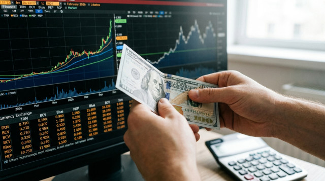 Una mano sosteniendo un billete de cien dólares frente a una pantalla con gráficos financieros y una calculadora, representando el análisis del tipo de cambio